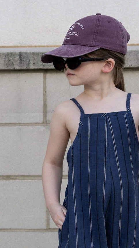 Close-up of girls overalls sewing pattern with adjustable metal strap hardware, gathered elastic back, navy pinstripe fabric, and oversized styling.